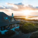 Une charmante maison en bord de mer en pierre avec un toit en bardeaux de cèdre et une cheminée, située sur une côte herbeuse au lever du soleil, face à l'océan Atlantique.