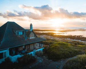 Une charmante maison en bord de mer en pierre avec un toit en bardeaux de cèdre et une cheminée, située sur une côte herbeuse au lever du soleil, face à l'océan Atlantique.