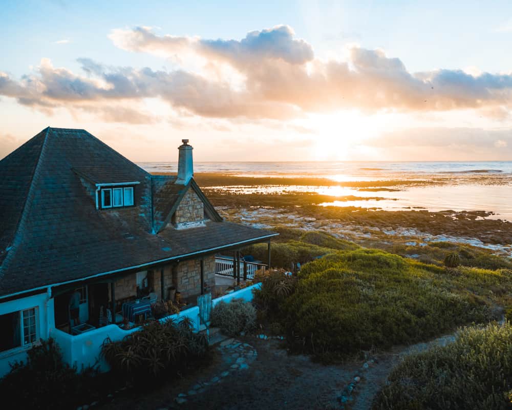 Une charmante maison en bord de mer en pierre avec un toit en bardeaux de cèdre et une cheminée, située sur une côte herbeuse au lever du soleil, face à l'océan Atlantique.